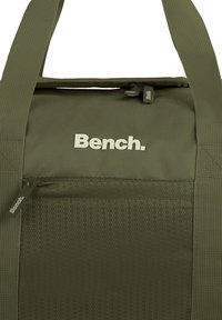 Groene stoffen draagtas met een hexagonaal patroon, logo in lichtgrijs, bovenste ritsluiting en verstelbare schouderband.