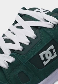 Σκούρο πράσινο αθλητικό παπούτσι με λευκά κορδόνια σφιχτά δεμένα και το λογότυπο DC Shoes στο πλάι και στη γλώσσα του παπουτσιού.
