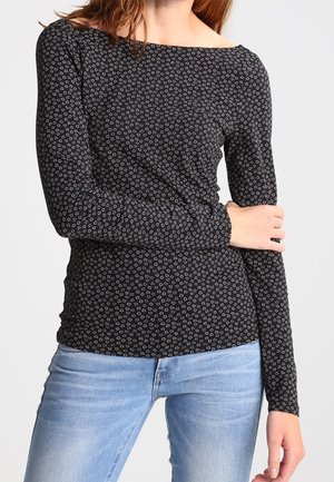 Femme portant un t-shirt noir à manches longues avec de petits motifs de feuilles blanches et un jean bleu clair, debout devant un fond clair uni.