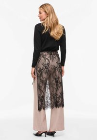 Jupe à superposition en dentelle noire avec une finition transparente, associée à un pantalon ample beige clair. La dentelle présente des motifs floraux complexes et des bords en feston.