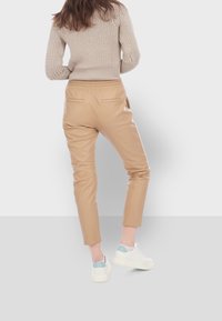 Světle hnědé kožené kalhoty s tapered střihem a elastickým pasem, vybavené dvěma zadními kapsami. Nosič má na sobě bílé tenisky a béžový pletený svetr.