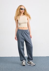 Top sin mangas beige, pantalones de chándal grises con cinturilla elástica y gafas de sol negras. Los zapatos presentan detalles metálicos y un diseño estampado.