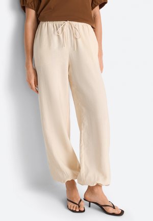Donna che indossa pantaloni beige larghi con vita elastica e coulisse, abbinati a sandali neri con tacco e top marrone.