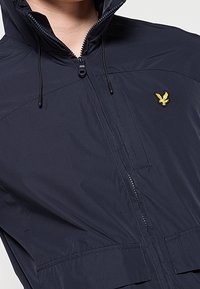 Personne portant une veste bleu marine foncé zippée avec un col relevé et un petit logo d'oiseau jaune sur la poitrine.