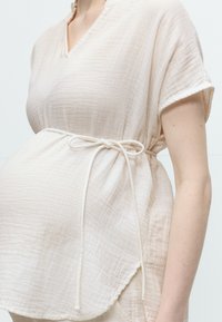 Ανοιχτό μπεζ υφασμάτινο τοπ για εγκύους με κοντά μανίκια και λεπτή ζώνη που δένει πάνω από την φουσκωμένη κοιλιά.