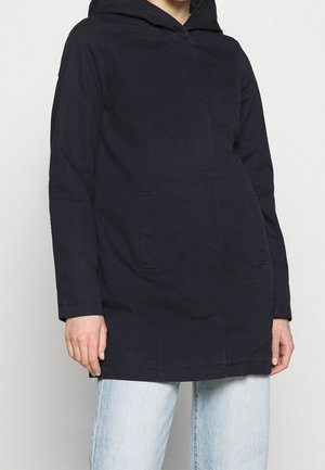 Personne portant une veste noire à capuche avec poches avant et un jean bleu clair, debout devant un fond clair uni.