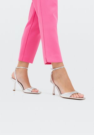 Pieds portant des sandales argentées à talons hauts avec brides à la cheville, associées à un pantalon court rose vif sur un fond uni.