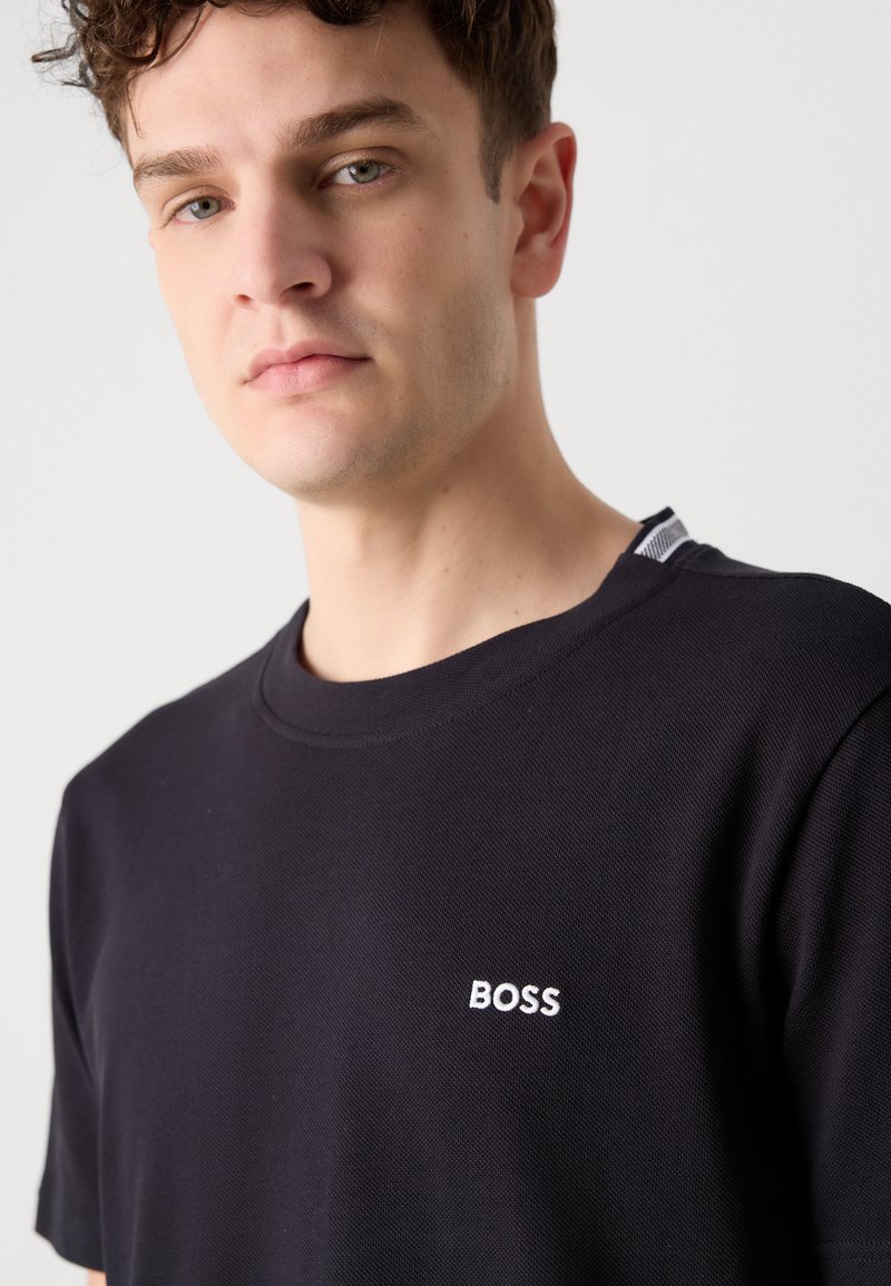 T-shirt noir à manches courtes en tissu texturé, avec un logo "BOSS" brodé en blanc sur le côté gauche de la poitrine. Détail accentué au niveau du col.