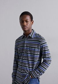 Πουκάμισο με κουμπιά και σχέδιο ζιγκ-ζαγκ σε πράσινο, μπλε και μαύρο. Φτιαγμένο από μαλακό ύφασμα, με μύτη κολάρο και μακριά μανίκια.