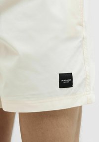 Costumi da bagno beige chiaro con una texture liscia, dotati di un'etichetta nera con testo bianco sull'orlo. Le cuciture sono visibili lungo i bordi.