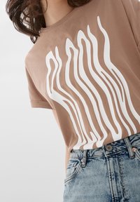 Hnědé bavlněné tričko s bílým vzorem vln; kulatý výstřih; doplněno světemodrými džínovými kraťasy s vybledlou texturou a kovovým knoflíkem.