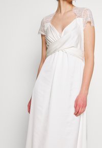 Robe blanche avec un décolleté en V profond, un corsage froncé et une ceinture en satin. Comprend des manches courtes en dentelle et une jupe fluide en tissu doux.