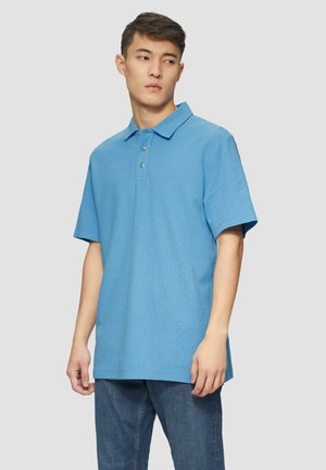 Camisa polo de color azul claro con mangas cortas, tapeta de tres botones y tejido texturizado. Combinada con jeans de color azul oscuro, diseño minimalista.