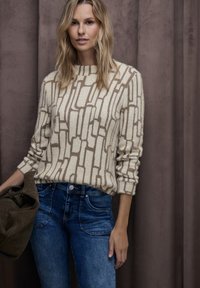 Beige tröja med ett geometriskt mönster i brunt, kombinerad med blå jeans i denim. Mjuk textur och rund halsringning. Modellen poserar mot en brun bakgrund.