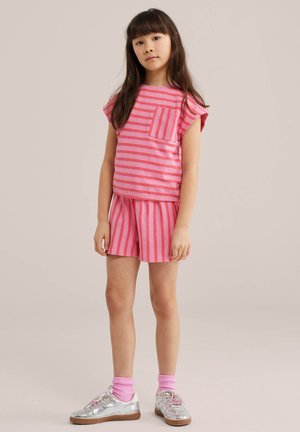 Mlada djevojka s dugom tamnom kosom u ružičastoj prugastoj majici i odgovarajućim kratkim hlačama, srebrnim tenisicama i ružičastim čarapama, stoji ispred obične pozadine.