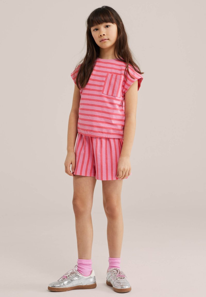 Jeune fille aux longs cheveux foncés portant un t-shirt rose rayé et un short assorti, des baskets argentées et des chaussettes roses, debout devant un fond uni.