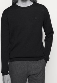 Svart stickad tröja med långa ärmar, rund hals och ribbade muddar. Har subtil broderad detalj på bröstet.