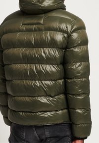 Chaqueta acolchada verde oliva con acabado brillante, con rayas acolchadas horizontales y capucha. Sin herrajes ni detalles visibles.