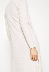 Vestido acanalado de color beige claro con mangas largas y una abertura lateral, que muestra una textura suave y un ajuste relajado.