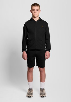 Teenagedreng stående iført sort lynlåshoodie og shorts, hvide sokker og beige sneakers mod en ensartet grå baggrund.