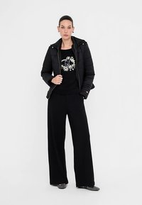 Zwarte pufferjas met ritssluiting, gecombineerd met een zwarte sweatshirt met bloemenprint en wijde zwarte broek. Grijze ballerina's.