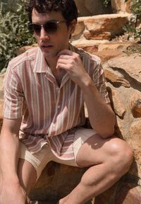 Gestreiftes Kurzarmhemd in Hellrosa und Weiß, aus strukturiertem Stoff. Kombiniert mit khakifarbenen Shorts, Modell sitzt vor steinfarbenem Hintergrund.