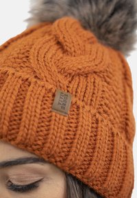 Berretto lavorato a maglia arancione con un motivo spesso e testurizzato e un pompon in pelliccia sintetica sulla parte superiore. Presenta una piccola etichetta in pelle sul lato.