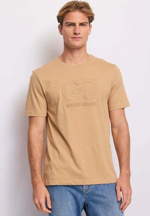 Giovane uomo che indossa una t-shirt beige Gaudi Jeans e jeans blu, in piedi davanti a uno sfondo chiaro e uniforme.