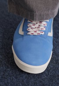 Sapatilha de camurça azul com detalhes brancos e atacadores vermelhos estampados, apresentando um upper liso e sola de borracha, parcialmente exibida contra um fundo azul.