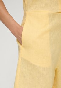 Geel linnen jumpsuit met een zichtbare zak, lichte textuur en subtiel patroon. Naaddetail zichtbaar aan de zijkant.