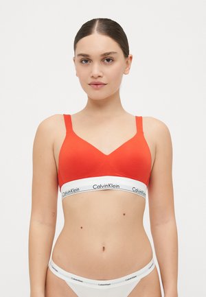 Punane puuvillane bralette V-kujulise kaelusega, laiade sangadega ja valge logoga elastse ribaga. Koos valgete bikiinipükstega.