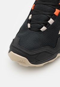 Gros plan d'une basket noire Adidas avec une semelle beige, une tige textile et des lacets noirs passés à travers des œillets beiges et orange.