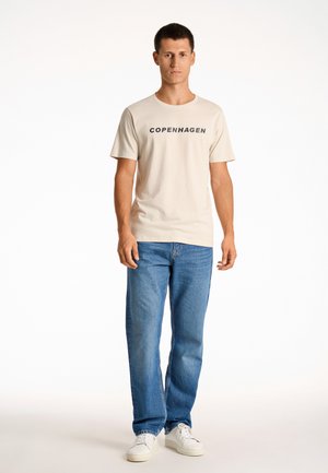 Camiseta de algodón color beige claro con "COPENHAGUE" impreso en negro. Combinada con jeans de mezclilla azules y zapatillas blancas. Corte y estilo casual.