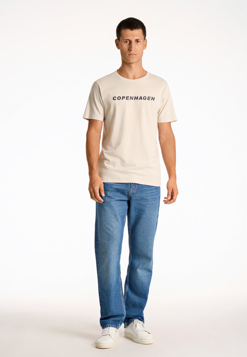 Camiseta de algodón color beige claro con "COPENHAGUE" impreso en negro. Combinada con jeans de mezclilla azules y zapatillas blancas. Corte y estilo casual.