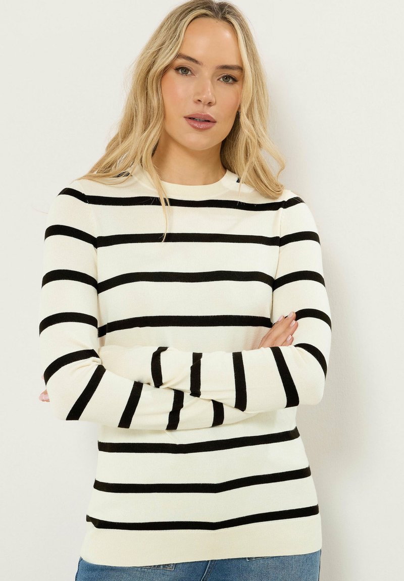 Mujer rubia con el cabello suelto, vistiendo un suéter blanco con rayas horizontales negras, con los brazos cruzados, posando frente a un fondo liso y claro.