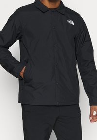 Homme portant une veste noire à boutons de North Face avec les mains dans les poches devant un fond uni.