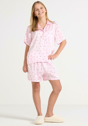 Smilende pige stående med hånd på hoften iført lyserødt blomsterprintet short-sleeve pyjamasæt og cremefarvede hjemmesko på hvid baggrund.