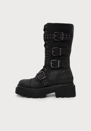 Botte de combat noire à mi-mollet, ornée de clous argentés, de plusieurs brides à boucles, de crochets pour les lacets et d’une épaisse semelle plateforme crantée.