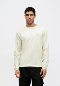 Κρεμ φούτερ με μακριά μανίκια από απαλό υλικό, με στρογγυλή λαιμόκοψη και μικρό χρυσό λογότυπο στο αριστερό στ�ήθος. Φορεμένο με σκούρο παντελόνι.