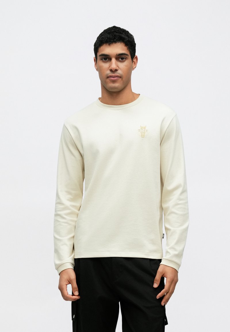 Κρεμ φούτερ με μακριά μανίκια από απαλό υλικό, με στρογγυλή λαιμόκοψη και μικρό χρυσό λογότυπο στο αριστερό στήθος. Φορεμένο με σκούρο παντελόνι.