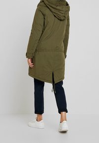 Olivgrön parka jacka med huva, med en längre bakdel, justerbart midjeband och sidofickor. Bärs med upprullade navybyxor och vita sneakers.