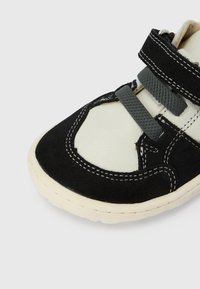 Μαύρο παπούτσι από σουέντ και λευκό δέρμα με γκρι λεπτομέρειες, χαρακτηριστικό στρογγυλή μύτη, ραμμένη διακόσμηση και δύο ελαστικούς ιμάντες για κλείσιμο.