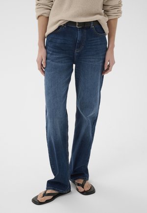 Oseba, oblečena v modre kavbojke srednjega pasu s črnim pasom, bež pulover in črne natikače s kvadratnim prstom, stoji na belem ozadju.