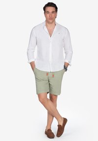 Camisa de lino blanca con botones, pantalones cortos verde claro con acentos naranjas y zapatos de lona marrones. Diseño sencillo, ajuste relajado.