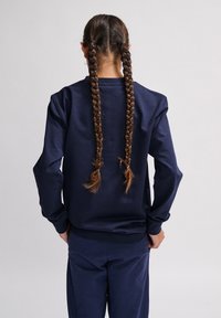 Felpa a maniche lunghe blu navy con collo rotondo, caratterizzata da una texture liscia e polsini a costine. Il modello ha due trecce raccolte in codini.