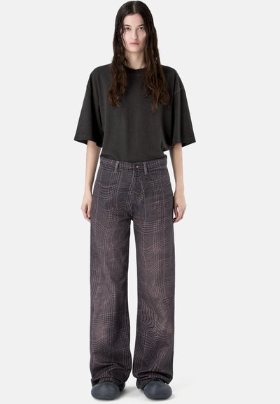 Frau mit langen dunklen Haaren, die ein lockeres schwarzes T-Shirt, graue Karo-Weitbein-Hosen und dunkle Slip-On-Schuhe trägt, stehend vor weißem Hintergrund.