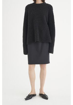Kvinde iført en sort tekstureret sweater, sort knælang nederdel og sorte spidse slip-on sko, stående mod en ensartet hvid baggrund.