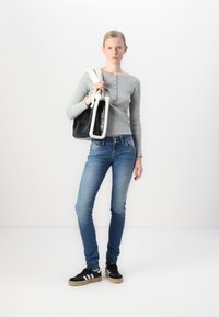 Haut à manches longues gris côtelé, jean skinny bleu et baskets noires avec des rayures blanches. Tient un sac noir avec des accents en fourrure blanche.