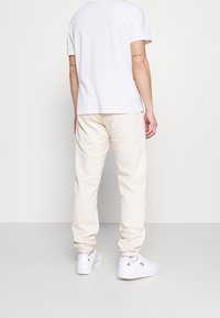 Man som bär vit t-shirt, beige joggerbyxor och vita Adidas-sneakers, stående med ryggen mot kameran framför en enfärgad ljus bakgrund.