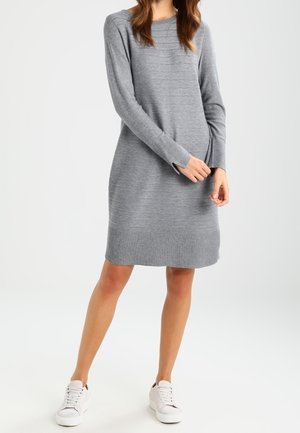 Donna che indossa un vestito maglione grigio a maniche lunghe fino al ginocchio e scarpe da ginnastica bianche, in piedi con una mano che tocca il polso dell'altra.